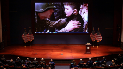 An image of a Ukrainian soldier and child plays on a projector screen in front of an audience of US lawmakers, with American flags on either side.