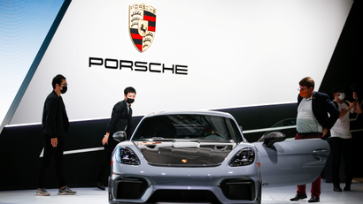 Three people gather around a Porsche car displayed at an auto show, with the brand's logo pictured above.