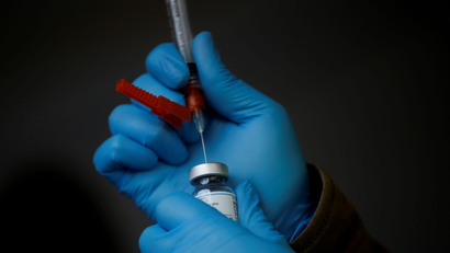 A person wearing blue latex gloves holds a syringe and vial of a Covid-19 vaccine