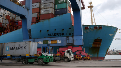 A Maersk ship docks at a port next to a truck carrying a Maersk container.