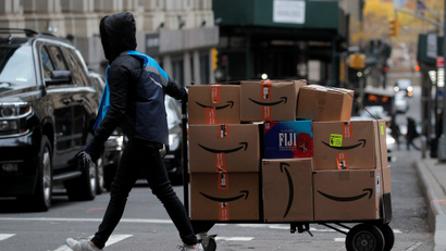 An Amazon delivery worker pulls a cart full of brown boxes for delivery outside an apartment building.