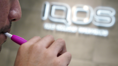 A person smoking a IQOS (I quit ordinary smoking) device in front of a pink sign that says IQOS
