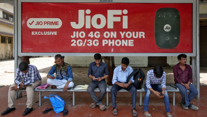 Commuters use their mobile phones as they wait at a bus stop with an advertisement of Reliance Industries' Jio telecoms unit, in Mumbai
