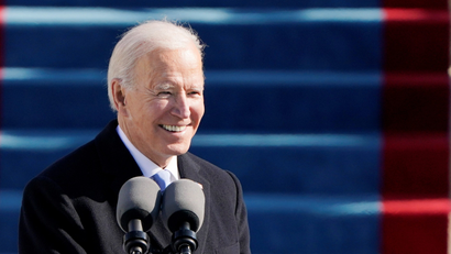 U.S. President Joe Biden smiles as he speaks into two large microphones during the 59th Presidential Inauguration in Washington, U.S., January 20, 2021. In the background is bright blue carpeted steps. Biden is wearing a black jacket, white shirt and grey tie.