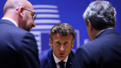 President Macron faces the camera head-on while speaking to two officials in the foreground, who are blurry. The background is a deep blue overlaid with a white design.