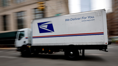 a white mail truck with a blue US Postal Service logo and blue and red stripe drives down a city street The background building is blurred to imply motion