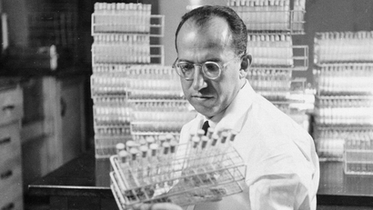 A black and white grainy image of Dr. Jonas Salk looking at vaccine viles.