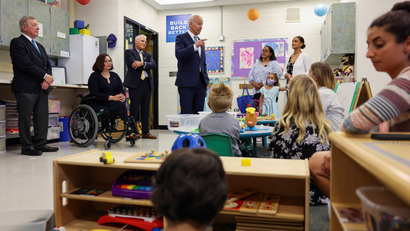 Senator Tammy Duckworth, seated in a wheelchair, President Joe Biden, and one other man, Clint Gabbard, stand in front of a classroom of children.