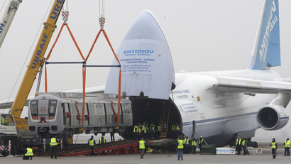 An An-124 sits on a runway. The plane's entire nose lifts up on a hinge to reveal its cavernous cargo hold. A crane heaves a train car into the opening, dwarfing the swarm of workers watching below.