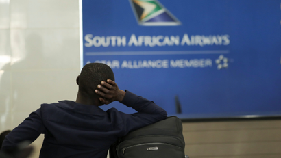 A seated passenger, with one arm resting on a bag, faces a sign for South African Airways at an airport