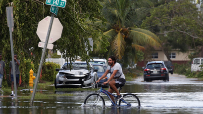 A man rides his bike down a flooded palm-tree lined street, through water that swallows up half of his tires.