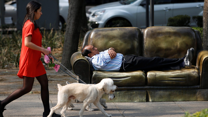 A man rests on a park bench in the shape of a sofa as a woman walks past with two dogs