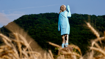 A crudely sculpted wooden sculpture of a woman wearing a light blue dress and waving with her left hand standing in a field with a green hill or mountain in the background