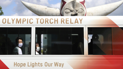 Two people wearing face masks look out the window of a bus that reads "Olympic Torch Relay"
