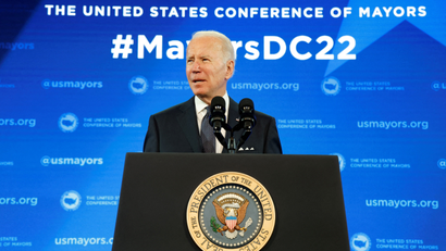 US president Joe Biden stands behind a podium with the official US presidential seal on it. Behind him is a graphic reading #MayorsDC22