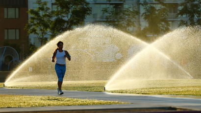 Sprinklers spray water onto grass as a jogger runs through a city park in San Diego, California