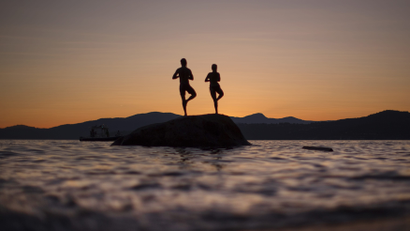 Two people stand on a rock facing the sunse