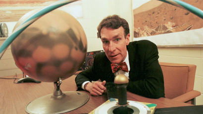 An image of Bill Nye sitting at a desk with a globe from the 90s.
