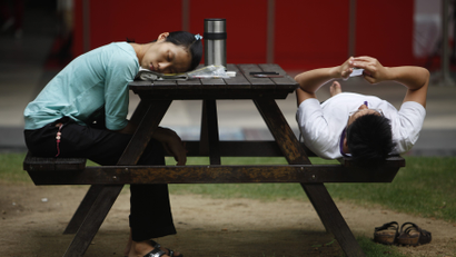 A woman sleeps with her head on a bench while another person checks their phone while lying down.
