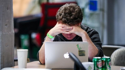 A stressed person holds their head in their hands while gazing at a laptop.