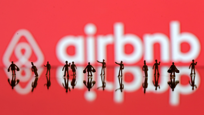 A 3D printed people's models are seen in front of a displayed Airbnb logo.