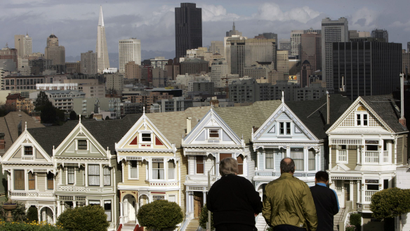 several Victorian homes in pastel shades of blue yellow and green are seen in front of the skyline of San Francisco