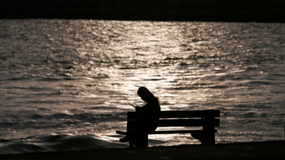 A woman sits alone on a bench while using her mobile phone as the sun sets at a beach.