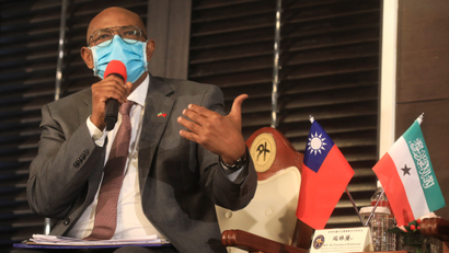 A man in a face mask holds a microphone and gestures. Beside him are two small flags.