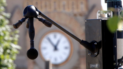 A black device with three microphones jutting out at angles from a central sphere, and one long microphone jutting out directly below are seen in the foreground in front of a clocktower in the background