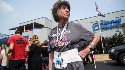 a woman clinic worker stands in front of a Planned Parenthood clinic. The woman has short hair and wears a grey t-shirt and lanyard.