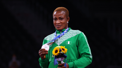 A picture of a Nigerian female wrestler holding her Silver medal from the 2020 Tokyo Olympics with her left hand. She looks delighted in her green tracksuit.