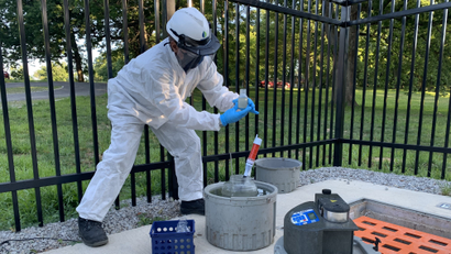 A person in a personal protective gear suit holds a sample of wastewater collected from the sewer below