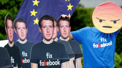 A protester wearing a shirt that reads "fix fakebook" and a mask that looks like Facebook's "angry reaction" stands next to cardboard cutouts depicting Facebook CEO Mark Zuckerberg