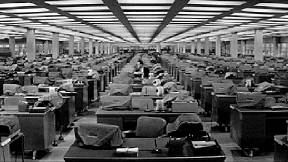 A black and white gif from an old movie of a man working alone in a large open-plan office.