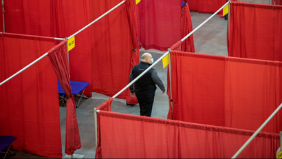 Red curtains make makeshift rooms in a large stadium. The beds are blue, and a single man with a mask on walks in between rows of curtains.