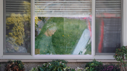An elderly woman with a mask on looking out of her nursing home window.