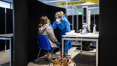 A woman wearing a blue gown, gloves, a face shield, and face mask tests a seated woman for covid-19 at Amsterdam's Schipol Airport.
