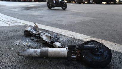 a burned out destroyed piece of a shared scooter lies on a concrete street. A moped can be seen in the background