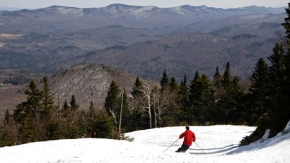 A man in a red coat skis down a snowy mountain, with forest and mountain range in the background