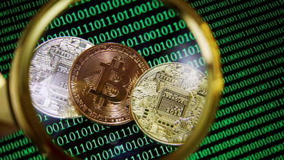 Representations of Bitcoin and other cryptocurrencies on a screen showing binary codes are seen through a magnifying glass