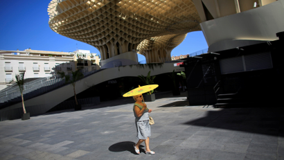 A woman stands alone in a public square on a sunny day with a yellow sun parasol. Behind her is a honeycomb like structure.