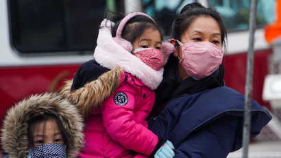 An Asian mother, wearing a light pink face mask and a dark blue winter coat, carries a young girl wearing a bright pink coat, pink earmuffs and a pink patterned facemask while standing next to an older young girl wearing a blue facemask. All three are watching Lunar New Year celebrations in New York City.