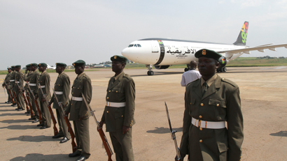 Men in uniform and guns stand at ease in a straight line. An airplane is in the background.