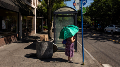 A woman stands with an umbrella to shade her from sun at a bus stop.