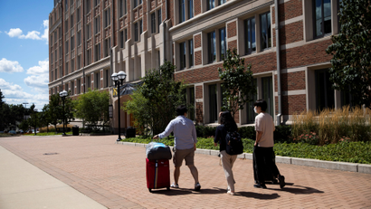 several students wheel suitcases down a brick path in front of a dormitory building