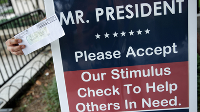 A hand outstretched holds a stimulus check in front of a sign that reads: "Mr. President, please accept our stimulus check to help others in need."