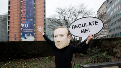 An activist wearing a paper mache Mark Zuckerberg mask holds a sign that says "Regulate me!"