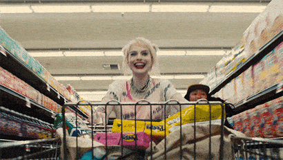 An animated gif of Harley Quinn running through a supermarket pushing a shopping cart while grinning maniacally.