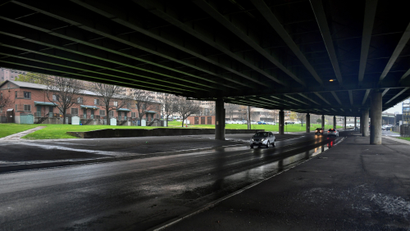 the view from a highway underpass with highway overheada sits above a road in the foreground, next to a row of apartments