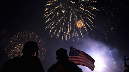 A silhouetted figure holds an American flag as fireworks burst in the night sky.
